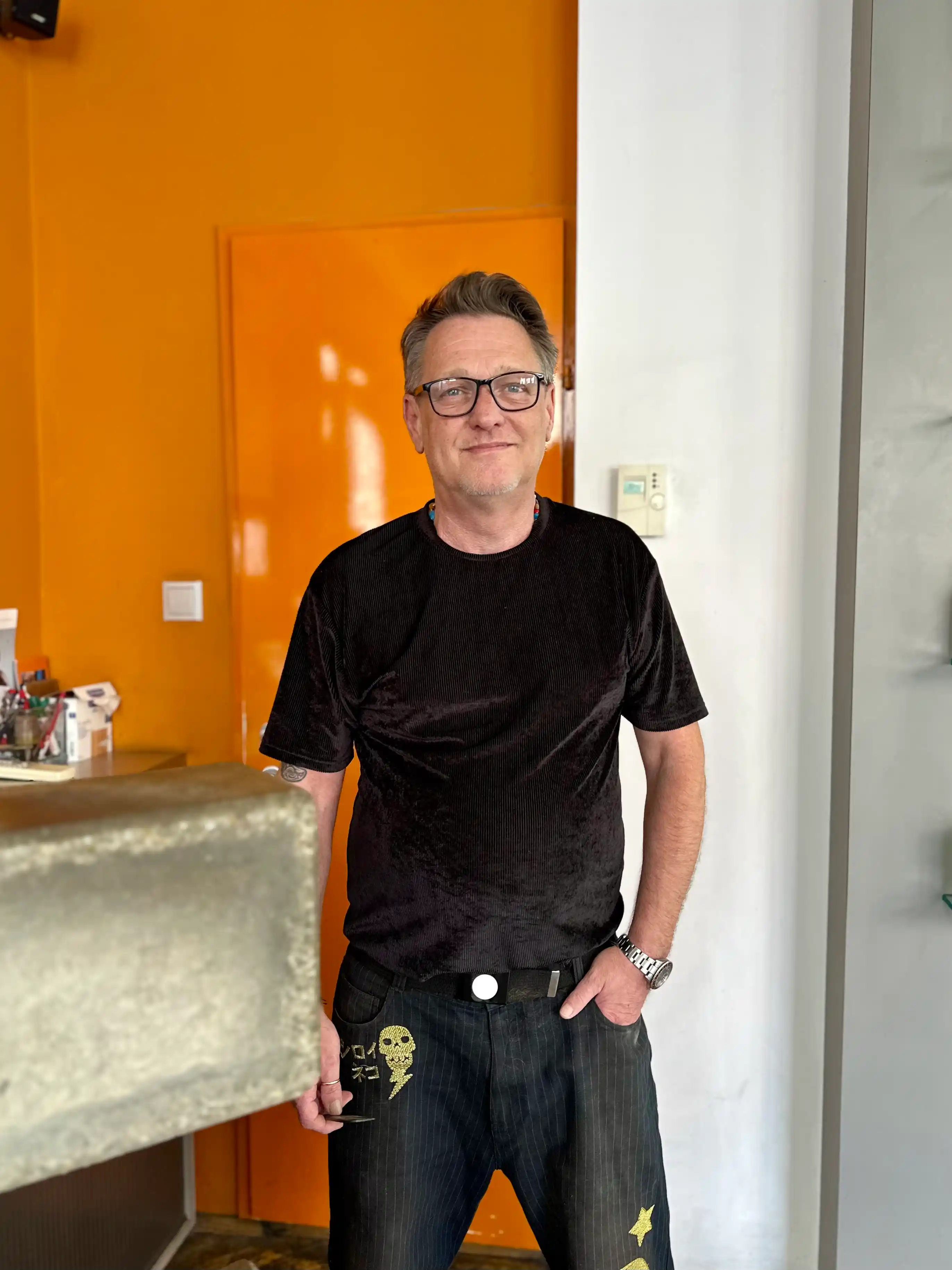 Manfred, Inhaber der HAIR-FACTORY, im Salon vor oranger Wand.