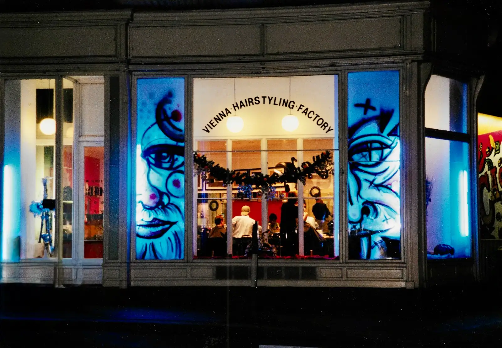 Das historische Schaufenster der HAIR-FACTORY bei Nacht — Clown-Graffiti und der Schriftzug Vienna Hairstyling Factory leuchten in der Dunkelheit.